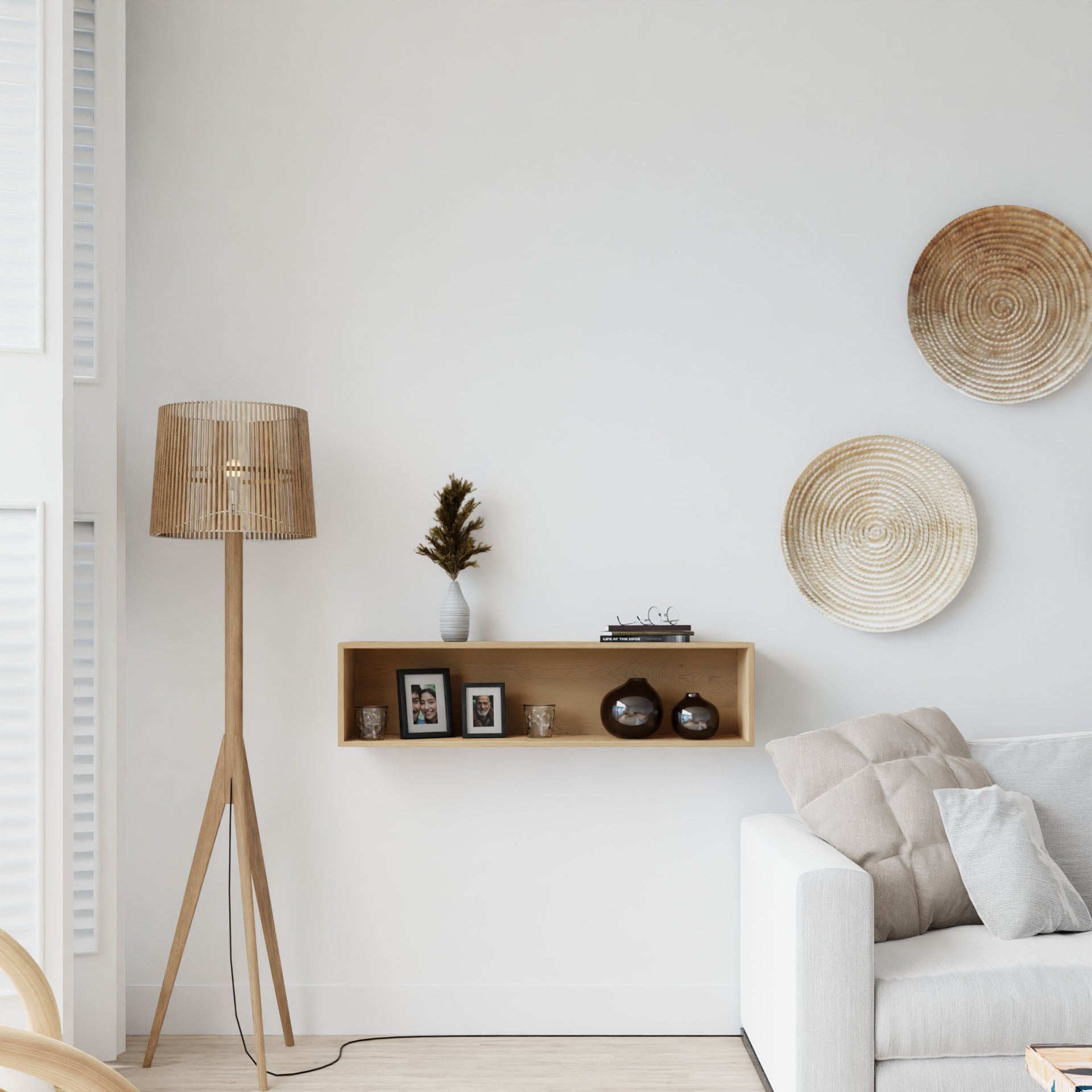 A minimalist living room with a neutral color palette showcases a floor lamp, Krovel's Floating Display Case in Maple adorned with small picture frames, decorative vases, and concentric-patterned plates on the wall. Partially visible on the right is a light-colored sofa.