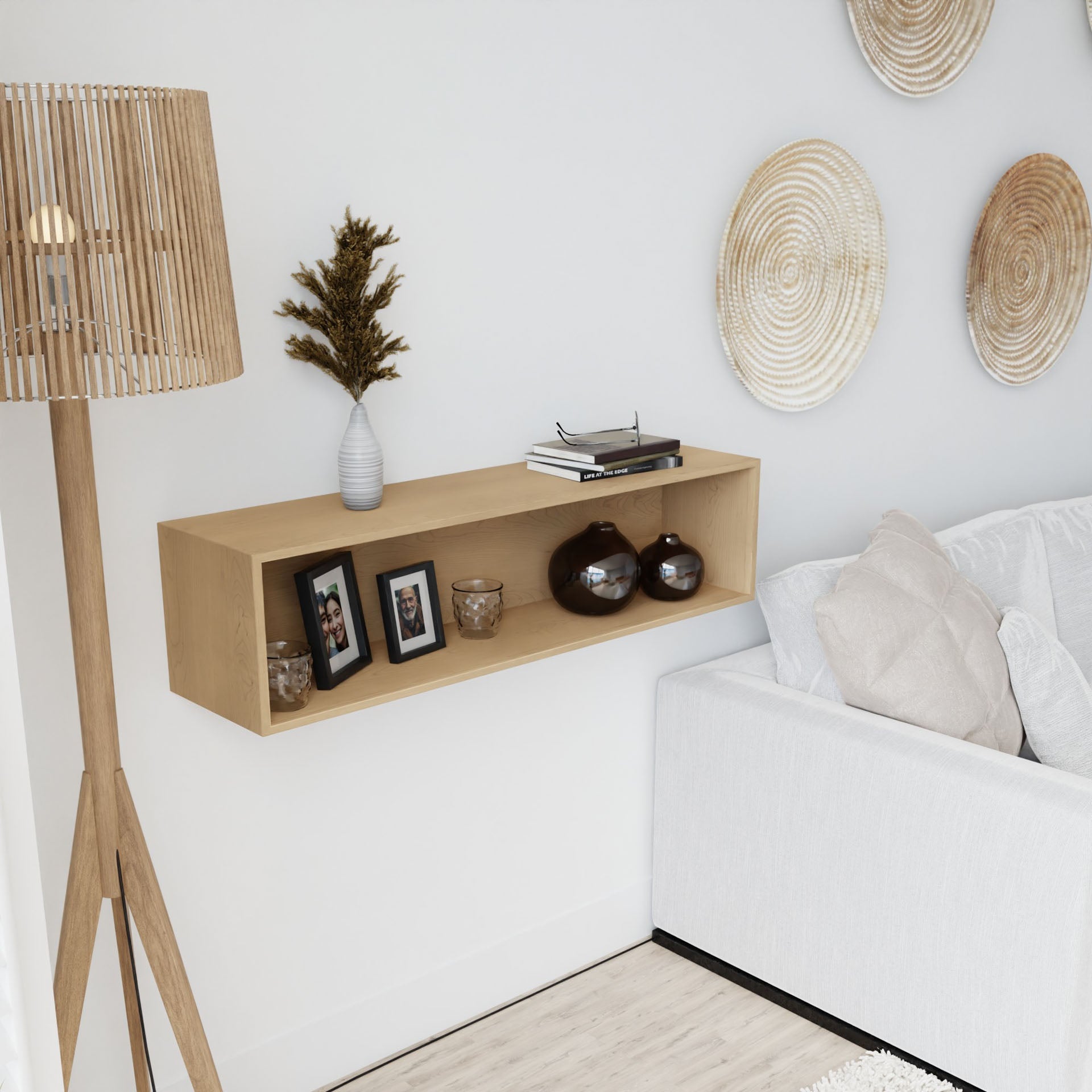 A modern living room showcases a white couch, a wooden floor lamp, and the elegant Krovel Floating Display Case in Maple. This display case holds framed photos, a vase with leafy branches, and decorative items. Woven baskets are displayed on the white wall above the couch, seamlessly supported by a discrete French cleat system.