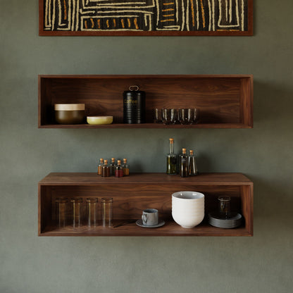 On a green wall, two Krovel's Floating Display Cases in Walnut showcase kitchen items. The top case features bowls, a black coffee canister, cups, and bottles, while the bottom case holds tall glasses, a white pot, a cup with saucer, and small bottles. Above them hangs a geometric-patterned art piece.