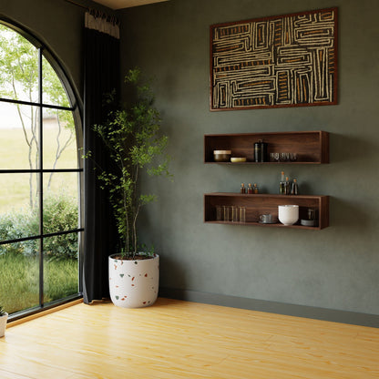 A minimalist room with a large arched window showcasing a scenic outdoor view features Krovel's Floating Display Case in Walnut that holds glassware and a white teapot. A tall plant in a white pot stands beside the display case, while geometric patterned artwork hangs above, all crafted from rich black walnut.