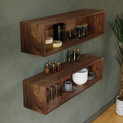 Two Krovel Floating Display Cases in Walnut adorn a green wall, showcasing a variety of items such as jars, mugs, a coffee container, oil bottles, and neatly stacked bowls. A potted plant is partially visible on the right.