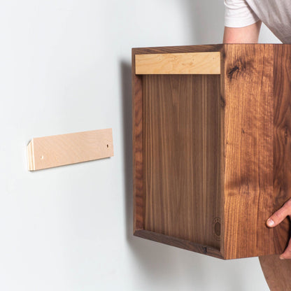 A person is expertly mounting a Krovel Floating Display Case in Walnut onto the wall using a French cleat system. The cleat, crafted from rich Black Walnut, is already attached to the wall, and the person aligns the display case for secure hanging, achieving a sleek look similar to floating shelves.