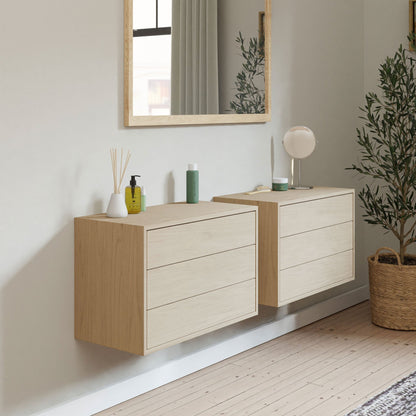 A minimalist room boasts two Krovel Floating Dressers in Maple with solid hard maple drawers and soft-close slides. A large mirror hangs above, reflecting natural light. A potted plant rests on the wooden floor, and toiletries are displayed on the floating dresser cabinets.