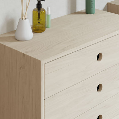 Krovel's Floating Dresser in Maple features a minimalist design with three drawers and circular cut-out handles. Topped with a yellow hand soap bottle, white vase with sticks, and small green spray bottle, it enhances the light ambience of the room.