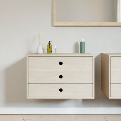 A minimalist bathroom features Krovel's Floating Dresser in Maple, a wall-mounted unit with three soft-close, black-handled drawers. Decorative items like a vase with reeds and toiletries adorn the dresser beneath a partially visible mirror.