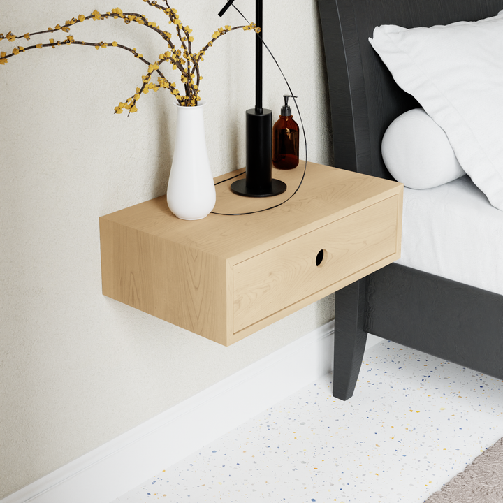 A handmade wall-mounted wooden bedside table with a drawer showcases Krovel Furniture Co.'s craftsmanship. It features a white vase with yellow flowers and a dark lamp. Next to the table is a gray bed adorned with white bedding and a round pillow, creating an inviting bedroom ambiance.