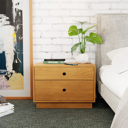 A Freestanding Nightstand in Cherry by Krovel with two drawers stands next to a gray upholstered bed. On the table are a potted plant, a book, and a smartphone. The background features a white brick wall and a large framed abstract painting.