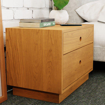 A Freestanding Nightstand in Cherry by Krovel with two drawers is placed beside a bed. On top, there are stacked books and potted greenery. The scene features a backdrop of a brick wall and a green carpeted floor, with all the solid hardwood elements contributing to a warm and inviting atmosphere.