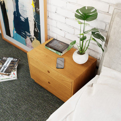 A Freestanding Nightstand in Cherry by Krovel with two drawers sits next to a bed. On top, there is a phone, a book titled "Escape," and a small potted plant with large green leaves. A modern painting leans against the white brick wall in the background.