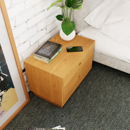 A Freestanding Nightstand in Cherry by Krovel stands beside a bed. Atop this solid hardwood furniture sit a plant in a white pot, a smartphone, and the book "Hard Truth." A framed picture leans against the white brick wall, while a carpet warmly covers the floor.