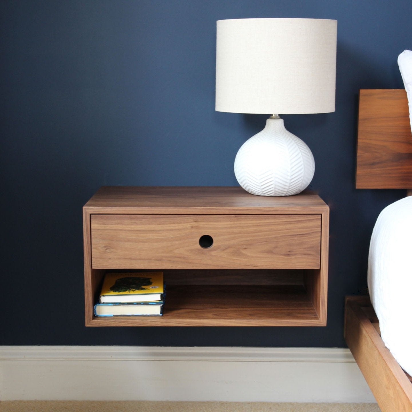 A modern handmade wooden floating nightstand from Krovel Furniture Co. against a dark blue wall. It features a drawer and an open shelf holding two books. On top sits a white textured lamp with a beige shade, beside a bed with crisp white linens.