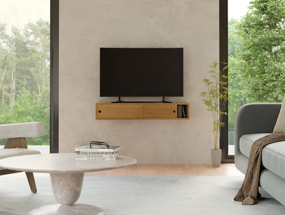 A modern living room with a minimalist design features a hand-finished wall-mounted flat-screen TV on a Krovel Media Console Table in White Oak, adorned with books. There’s a gray sofa, a round marble coffee table with magazines, and large windows revealing a green, leafy outdoor view.