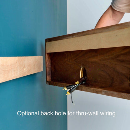 A person is installing a hand-finished wooden frame with a hole for wiring against a blue wall, complemented by an American Cherry bracket. Text on the image reads, "Optional back hole for thru-wall wiring." Product displayed: Media Console Table in White Oak by Krovel.