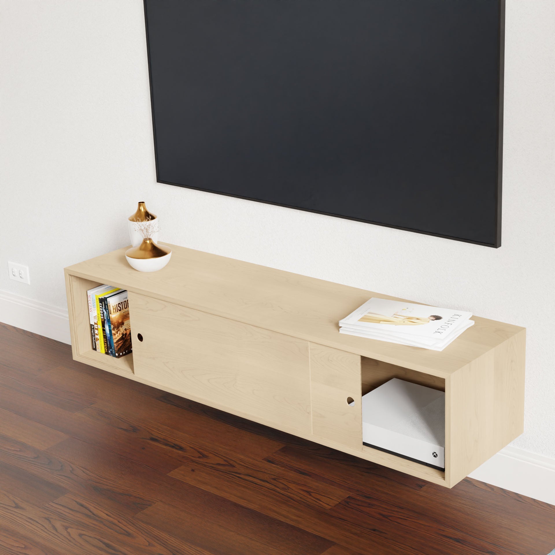 An Oversized Media Console Table in Maple by Krovel, hand-finished in a light tone inspired by American Cherry, includes side shelving adorned with books and decorative items. Above the floating XL console is a large, plain black screen mounted on a white wall, complemented by a floor of polished dark wood.