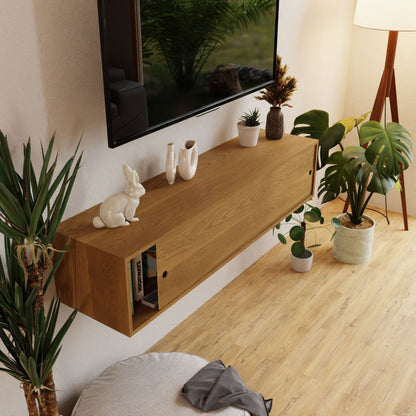 A contemporary living room showcases the Oversized Media Console Table by Krovel, crafted from White Oak. Above it is a mounted wooden shelf displaying decorative items such as a white rabbit figurine and vases. The area is adorned with various potted plants, while a TV is mounted above the shelf using a French cleat. A nearby lamp stands gracefully on the wooden floor.