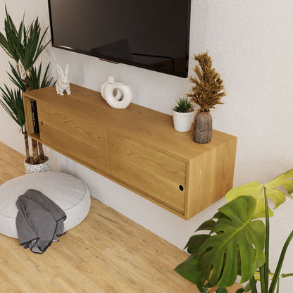 A contemporary living room arrangement showcases a Krovel Oversized Media Console Table in White Oak. Mounted above is a flat-screen TV, complemented by decorative elements such as a plant, ceramic bunny, and abstract sculpture adorning the console. A round cushion and a large leaf plant enhance the setup on the light wood floor.