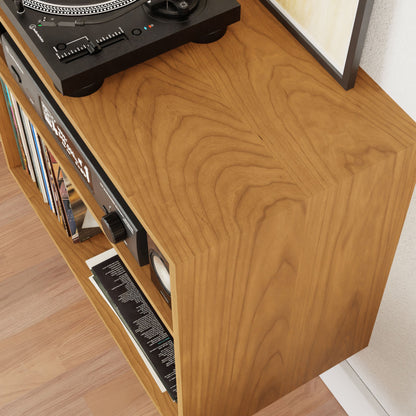 The Record and Stereo Storage Cabinet in Cherry by Krovel is a beautifully crafted wooden console table in a light brown finish, perfect for holding a vinyl record player on top. It features a side shelf for storing records and is made from solid American cherry, highlighting an eye-catching wood grain pattern that complements the light wooden floor it stands on.