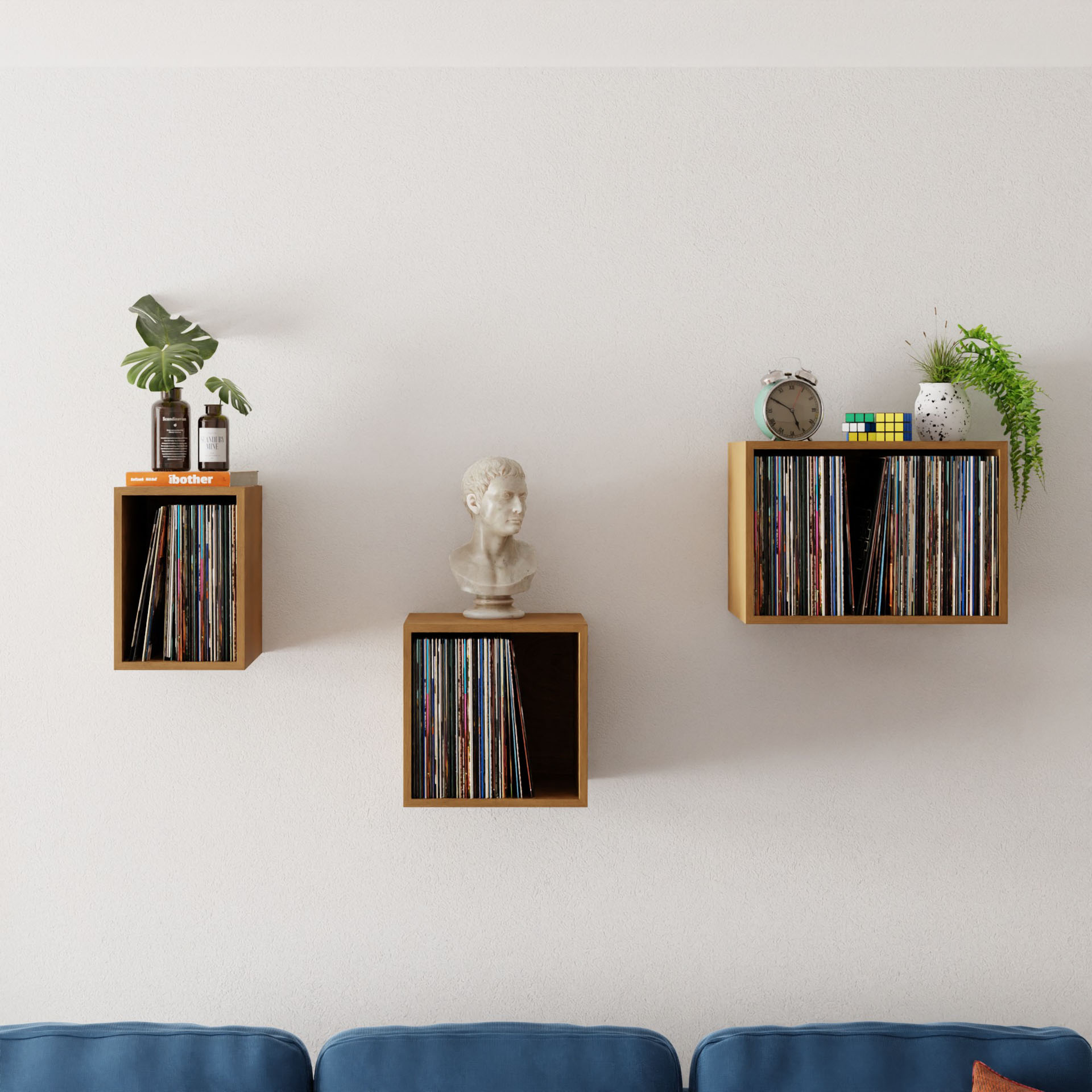 Krovel's Record Storage Shelves in Cherry stylishly display a vinyl collection, a plant, and decorative items. These shelves form an artistic arrangement above a blue sofa, beautifully contrasted against a crisp white wall.