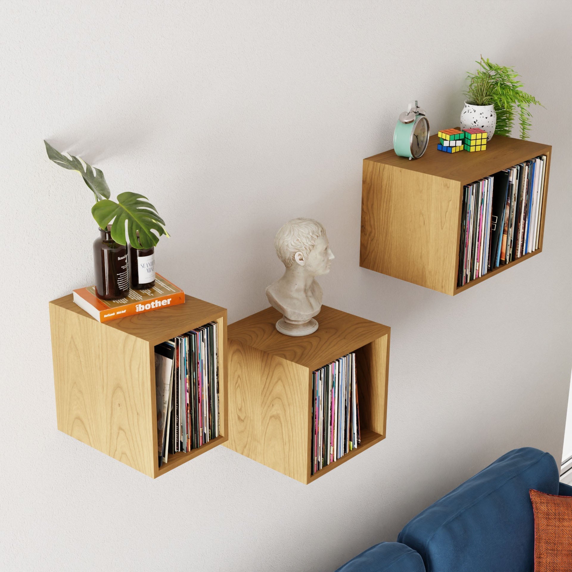 Three wall-mounted shelves, crafted by Krovel from luxurious Cherry wood, elegantly hold books, a potted plant, a globe, a Rubik's cube, and a bust sculpture. They beautifully display treasures above the blue couch. A model ship is proudly placed beside the Krovel Record Storage Shelves amidst an array of vinyl records.