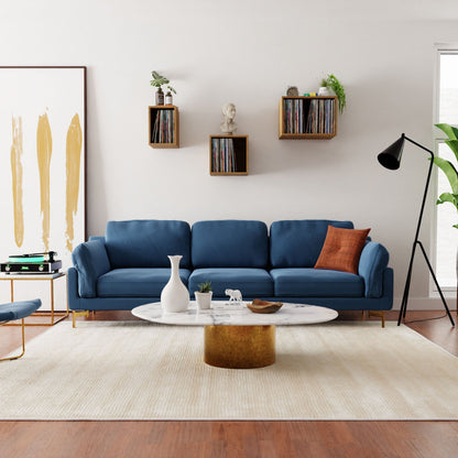 In a contemporary living room, a blue sofa is paired with a white round coffee table. A floor lamp stands close by, while floating record shelves by Krovel, made from Cherry wood, elegantly display a vinyl collection above the sofa. Leaning against the wall is a large abstract painting, and a plant rests beside the window.