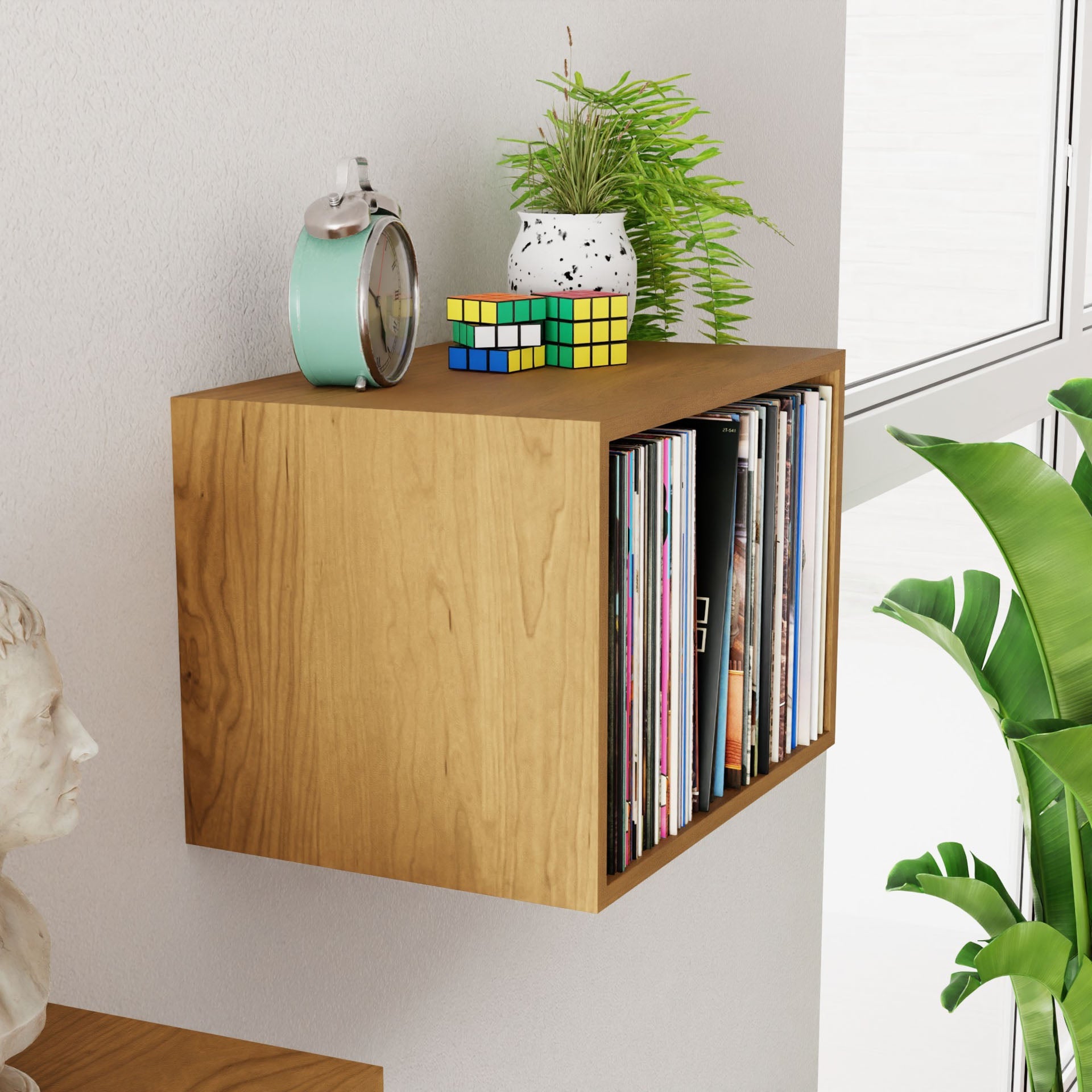 A Cherry wall-mounted shelf from Krovel serves as a perfect spot for displaying a vintage clock, a potted plant, and stacked Rubik's cubes. The side doubles as vinyl collection storage. Adjacent to the shelf are a small white statue and large green plant leaves, all beautifully lit by the natural light from the window.