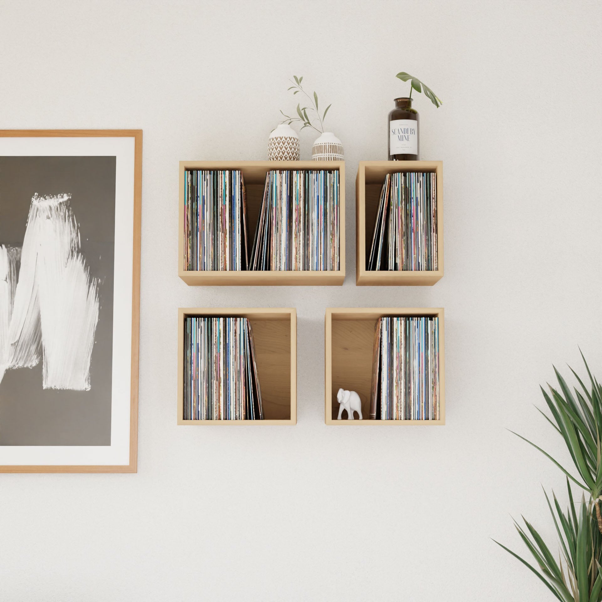 In a minimalist room, a framed abstract painting hangs on a white wall next to Krovel's Record Storage Shelves in Maple, housing a vinyl collection. Two small potted plants and a decorative item enhance the mid-century modern shelves, while a leafy plant adds greenery in the bottom right corner.