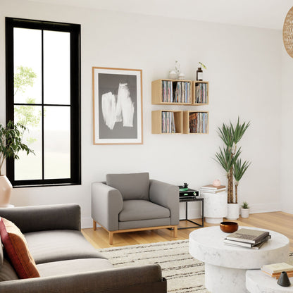 A minimalist living room featuring a gray armchair, sofa, and round coffee table exudes mid-century modern elegance. A large window frames an abstract painting, while Krovel's Record Storage Shelves in Maple beautifully display books and a vinyl collection. A small side table with decor and a potted plant complete the neutral palette.