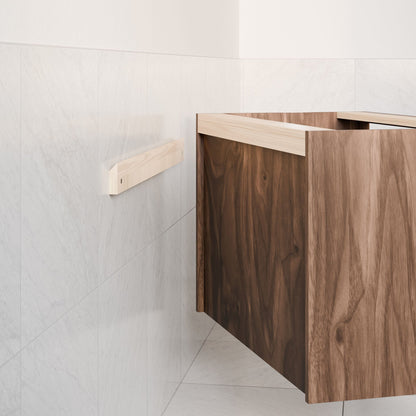 A Single Door, Triple Drawer Vanity in Walnut by Krovel Furniture Co. with soft-close hinges is partially open against a smooth, light-tiled wall. To the left of the rich, dark hardwood veneer vanity, a wooden rail is mounted on the wall.