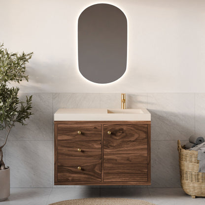 In a bathroom scene, a Krovel Furniture Co. Single Door, Triple Drawer Vanity in Walnut pairs with a white sink and brass fixtures beneath an oval backlit mirror. To the left is a potted plant; to the right, a wicker basket with rolled towels complements the neutral tile walls.