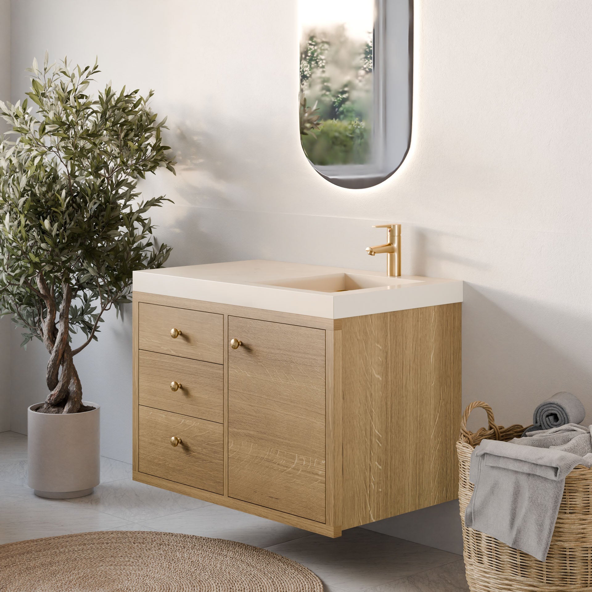 A minimalist bathroom features Krovel Furniture Co.'s Single Door, Triple Drawer Vanity in White Oak with gold handles, a white countertop, and a gold faucet. An oval mirror hangs above while a woven basket of towels and a potted plant adds charm to the serene space.