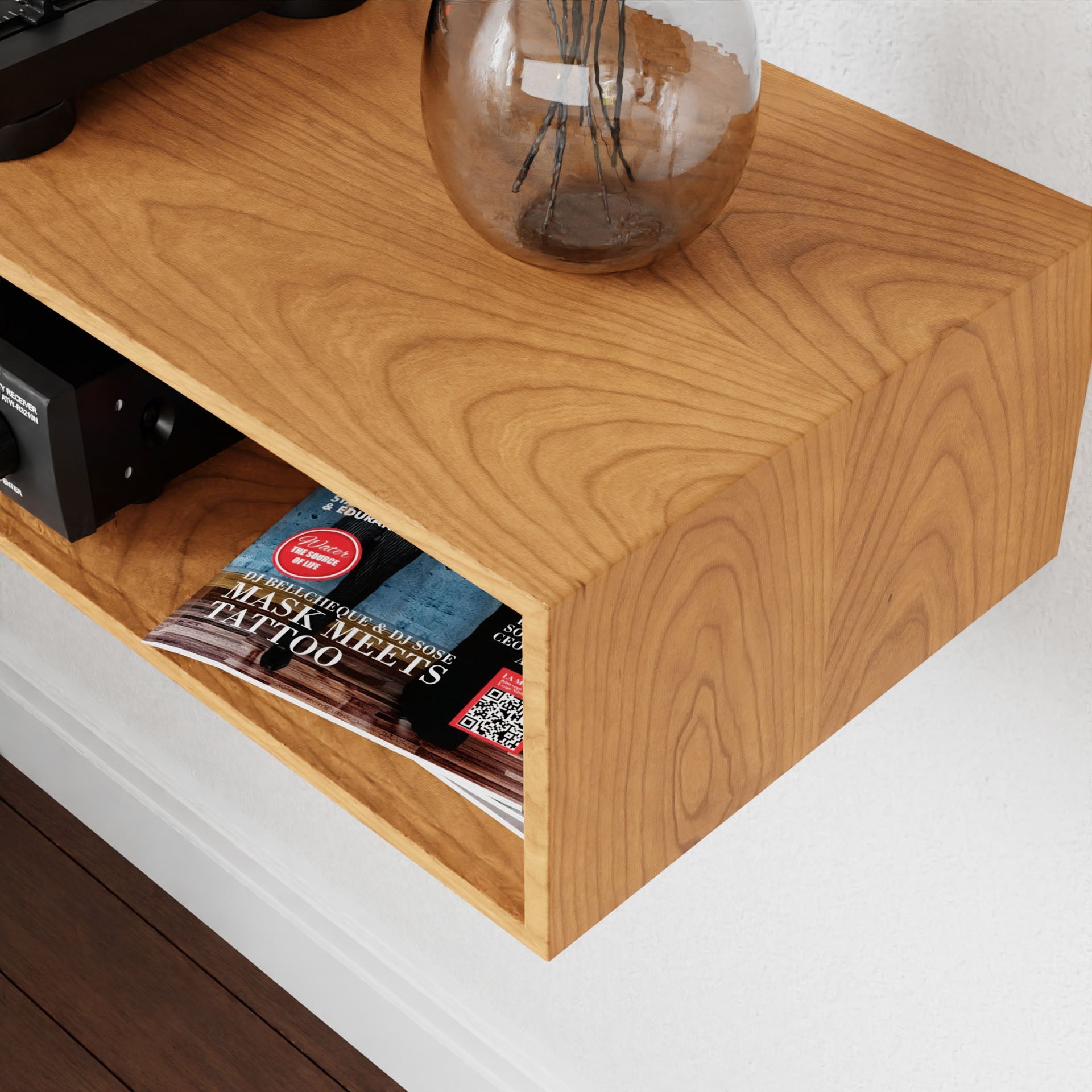 A Stereo Table in Cherry from Krovel, crafted from solid American Cherry, elegantly holds a glass vase with branches and a small electronic device. Below, a magazine with red and black cover art peeks out from the open storage space. The mid-century modern design is beautifully mounted on a white wall.