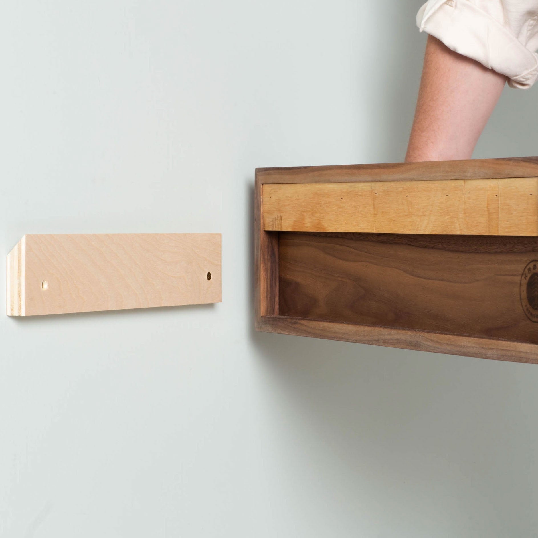 A person is installing a Krovel Stereo Table in Walnut on a wall using a mount. The person's arm is partially visible, and the table, with its hollow design reminiscent of mid-century modern style, slides effortlessly onto the mount. The wall is painted light green.