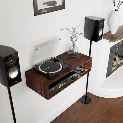 A modern living room showcases a mid-century modern aesthetic featuring the Krovel Stereo Table in Walnut, designed to mimic an elegant console table and accommodate a turntable and amplifier. Crafted from American Black Walnut, this setup is adorned with two standing speakers. Above the fireplace, a vase with branches adds charm, accompanied by a framed picture on the wall.