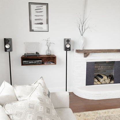 A modern living room with a neutral color scheme showcases a white brick fireplace and a cozy couch with patterned pillows. A mid-century modern Krovel Stereo Table in Walnut supports a record player and speaker, while artwork decorates the walls and an American Black Walnut vase with branches adds an elegant touch.