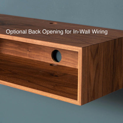 The Stereo Table in Walnut by Krovel, designed in a mid-century modern style, is crafted from American Black Walnut and includes an optional circular opening for seamless in-wall wiring. When mounted against a blue-green wall, it combines both style and functionality to enhance any room. The text reads "Optional Back Opening for In-Wall Wiring.